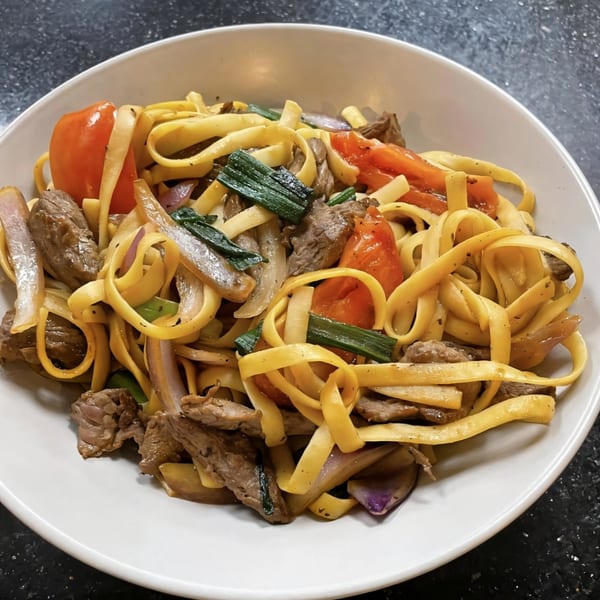Peruvian Lomo Saltado with fettuccine, beef, tomatoes, onions, and green onions in a white bowl.