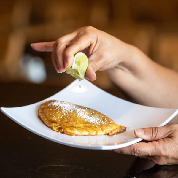 Hands squeezing lime juice onto a golden Peruvian empanada dusted with powdered sugar on a white plate.