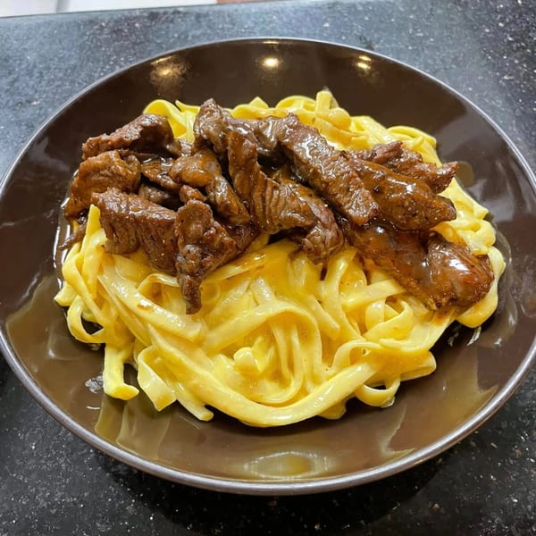 Peruvian Lomo Saltado with fettuccine Alfredo, featuring tender beef strips over creamy pasta in a brown bowl.